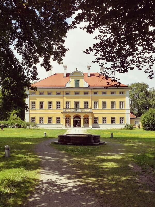 Blick auf die Frohnburg, ein kleines gelbes Schloss in einer sonnigen Parkanlage. Kieswege, Bäume, ein kleiner Brunnen