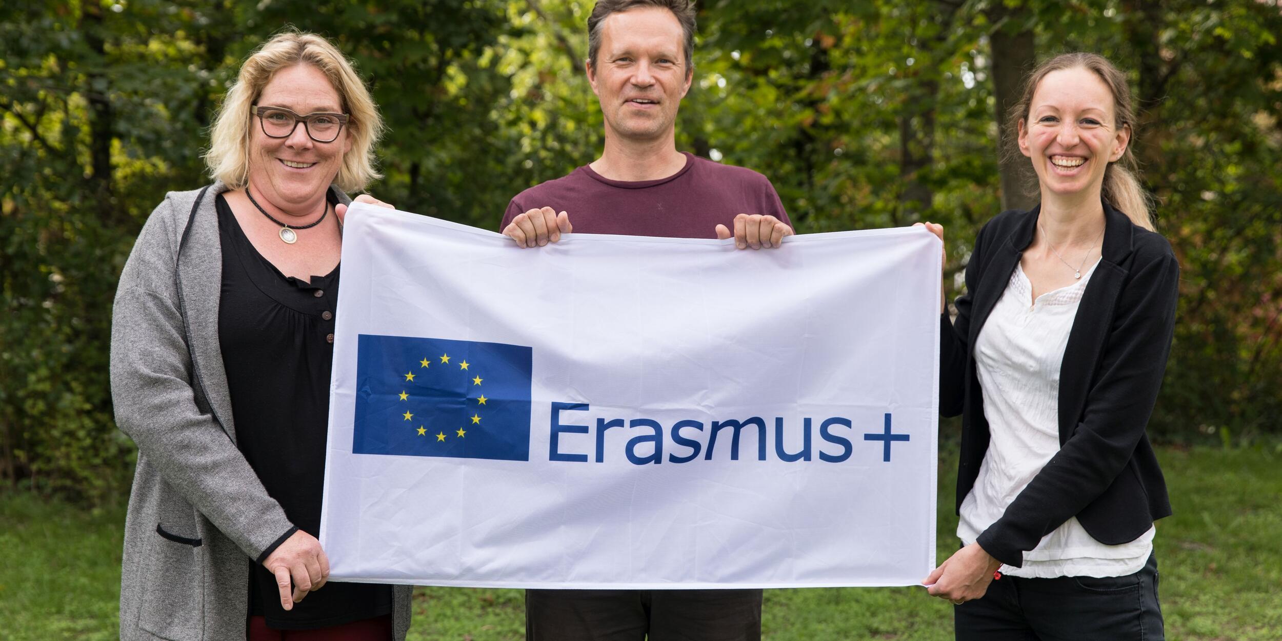 Drei Menschen auf einer Wiese, dahinter Wald, sie halten eine Erasmus-Flagge