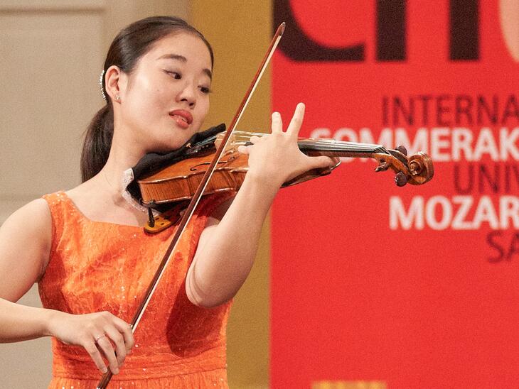 Asiatische Frau in orangem Kleid spielt Violine, roter Hintergrund | © Christian Schneider