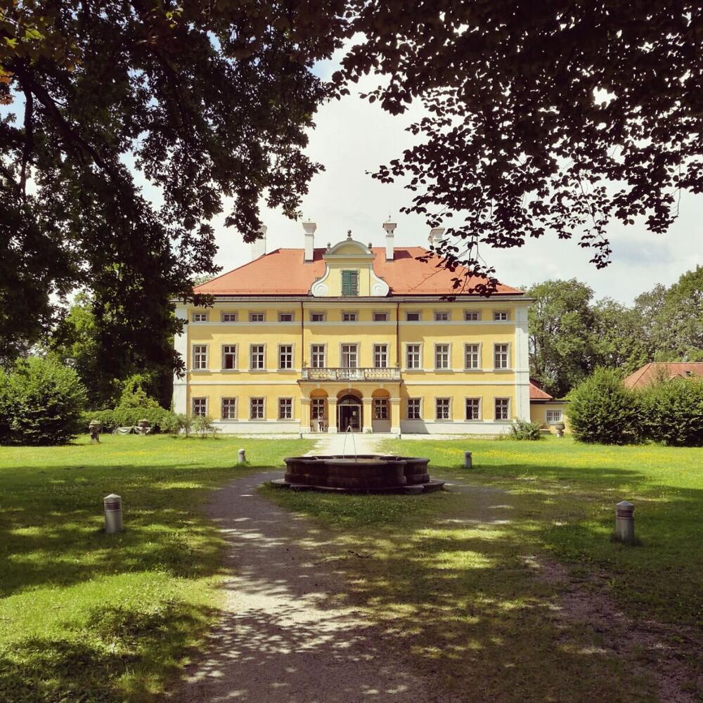 Blick auf die Frohnburg, ein kleines gelbes Schloss in einer sonnigen Parkanlage. Kieswege, Bäume, ein kleiner Brunnen