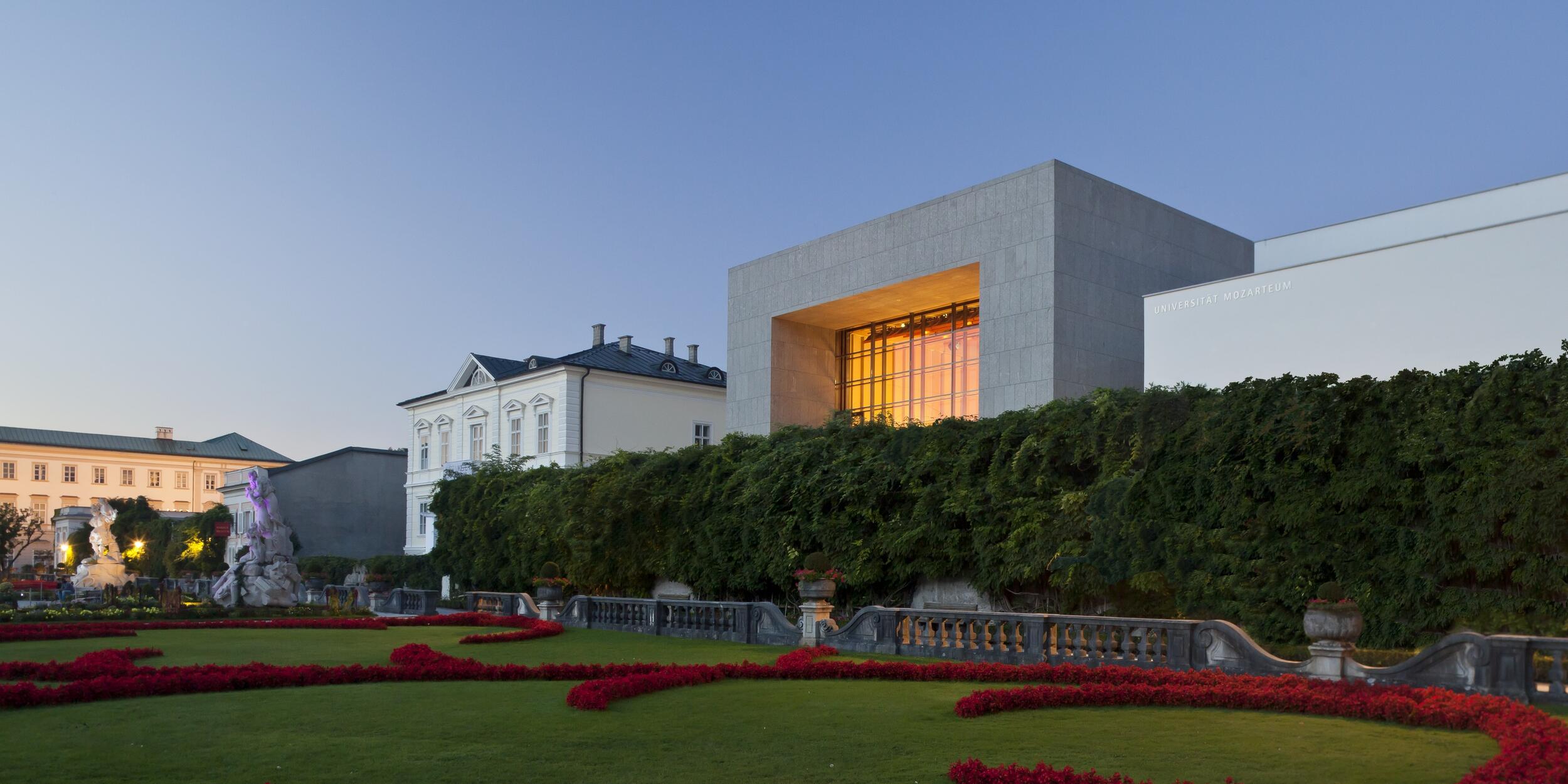 Blick auf den Solitär aus dem Mirabellgarten, die große Fensterfront leuchtet orange, Abendstimmung