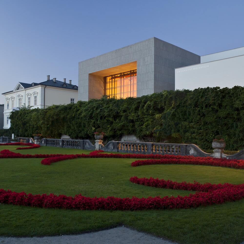 Blick auf den Solitär aus dem Mirabellgarten, die große Fensterfront leuchtet orange, Abendstimmung